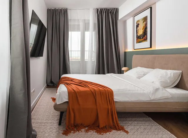 Bedroom with a double bed, an orange throw and grey curtains, at City of Mara, Timișoara
